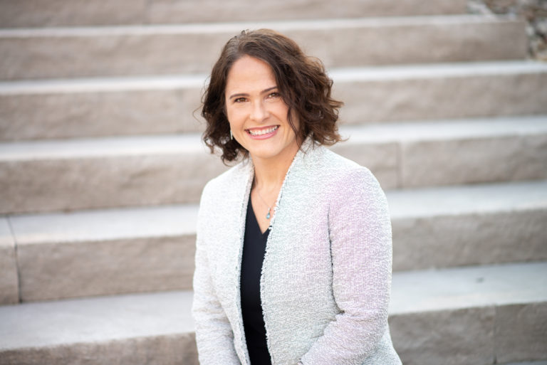 Professional woman with curly hair smiling while seated on stone steps, representing the Duckett Ladd Advisors team focused on dental practice financial services.