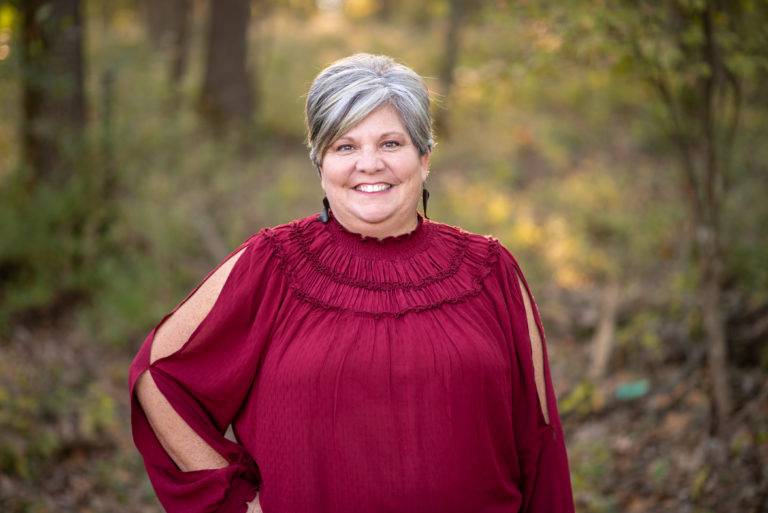 Angie Lippold smiling outdoors in a burgundy blouse, representing the team at Duckett Ladd Advisors, specializing in financial services for dental practices.