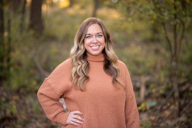 Smiling woman in an orange sweater standing outdoors in a natural setting, representing the team at Duckett Ladd Advisors, LLC, specializing in financial services for dental practices.