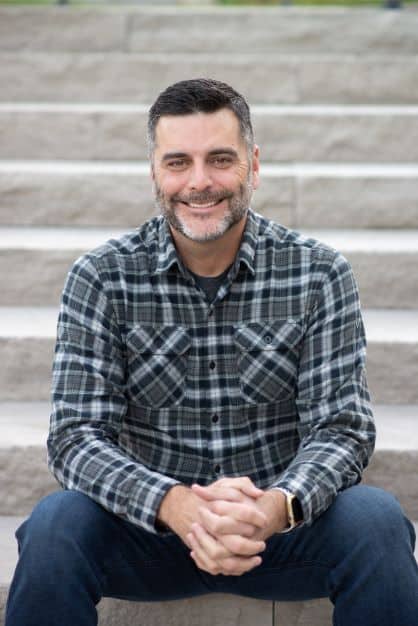 Smiling man in plaid shirt sitting on steps, representing the team at Duckett Ladd Advisors, LLC, specializing in financial services for dental practices.