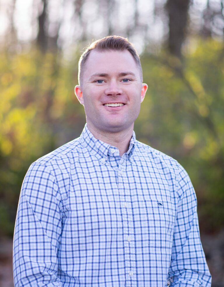 Caleb Gussner, team member at Duckett Ladd Advisors, smiling outdoors in a checked shirt, surrounded by greenery, representing expertise in financial services for dental practices.