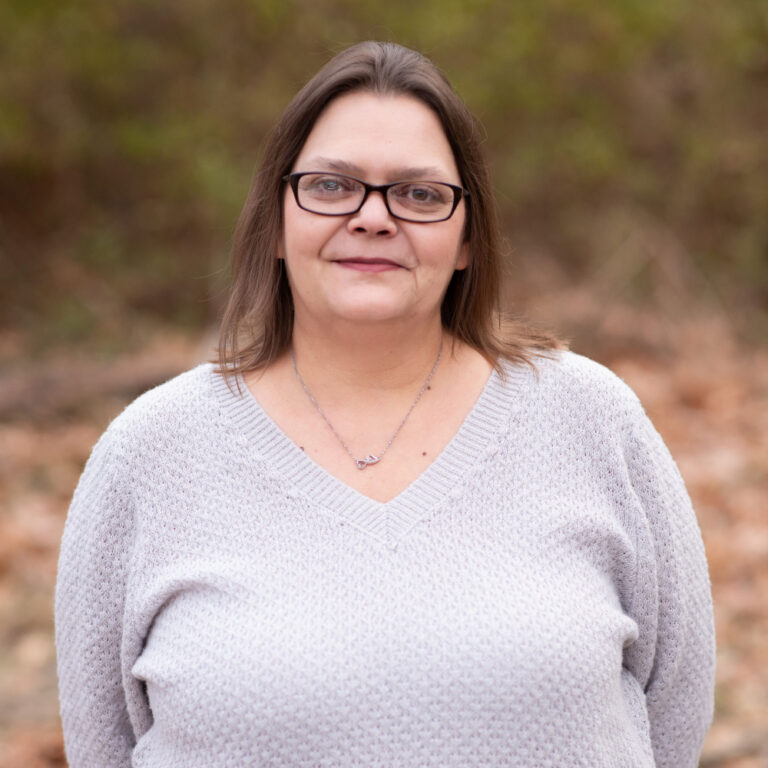 Alisha Vance, team member at Duckett Ladd Advisors, wearing a light gray sweater, smiling in an outdoor setting with blurred greenery in the background.